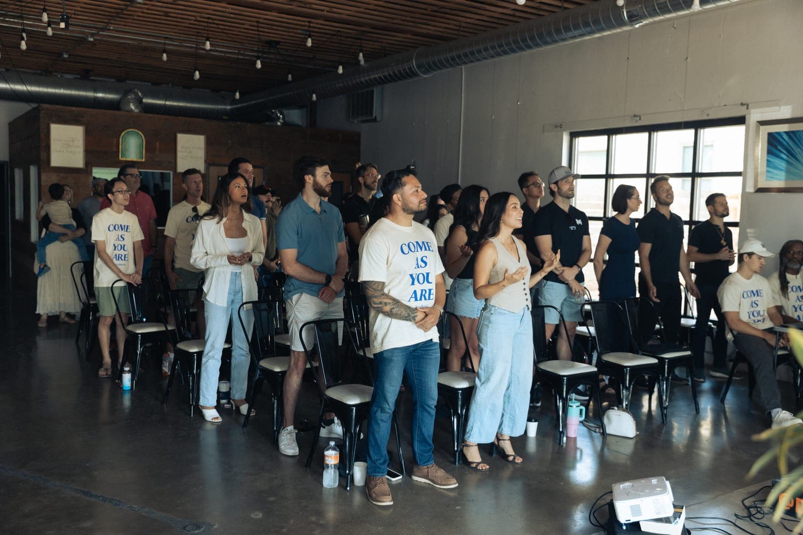 People stand during worship under string lights on a concrete floor, several in come as you are T-shirts.
