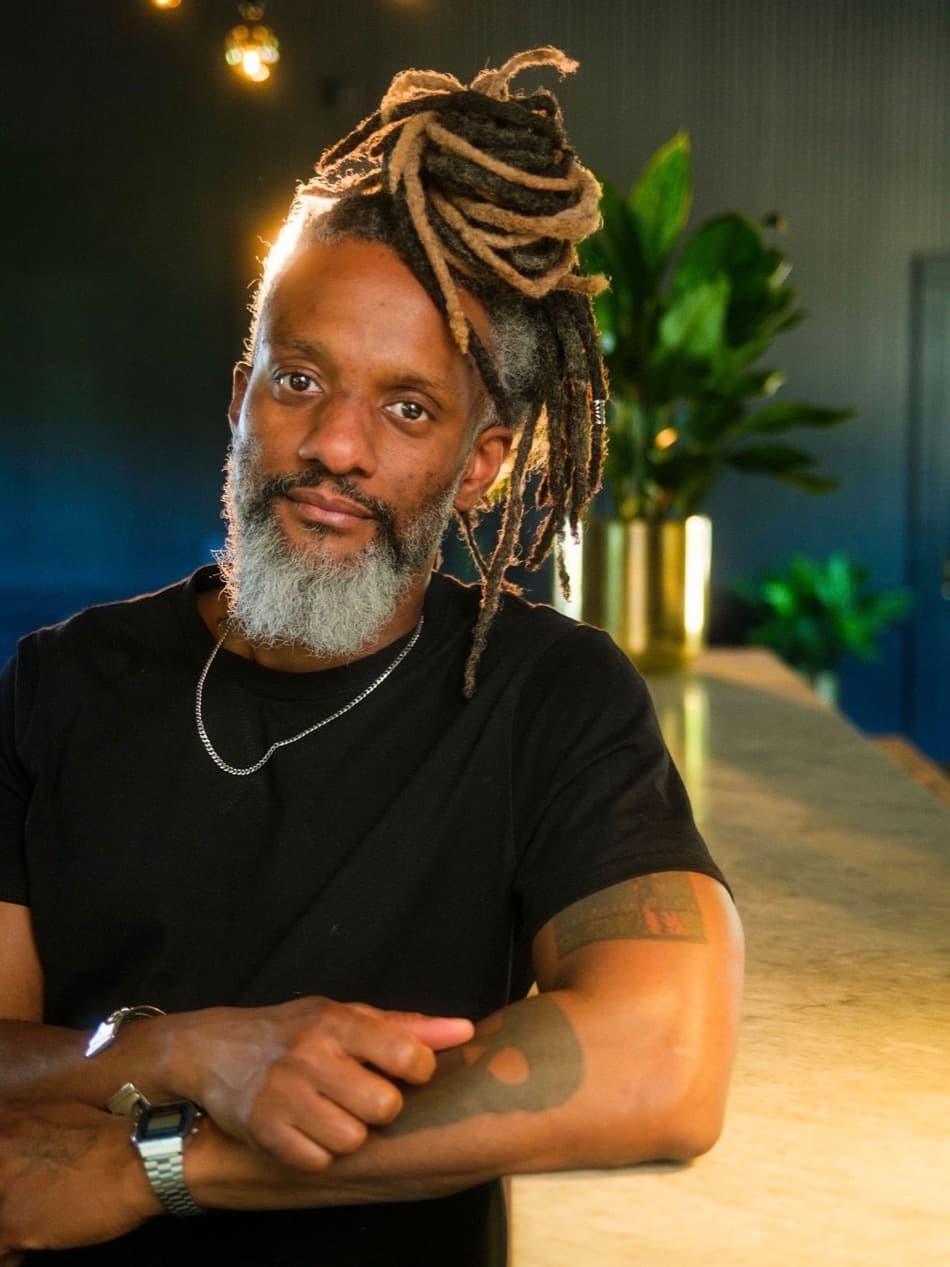 Khary Alexander in a black T-shirt, arms folded on a counter under warm pendant lights