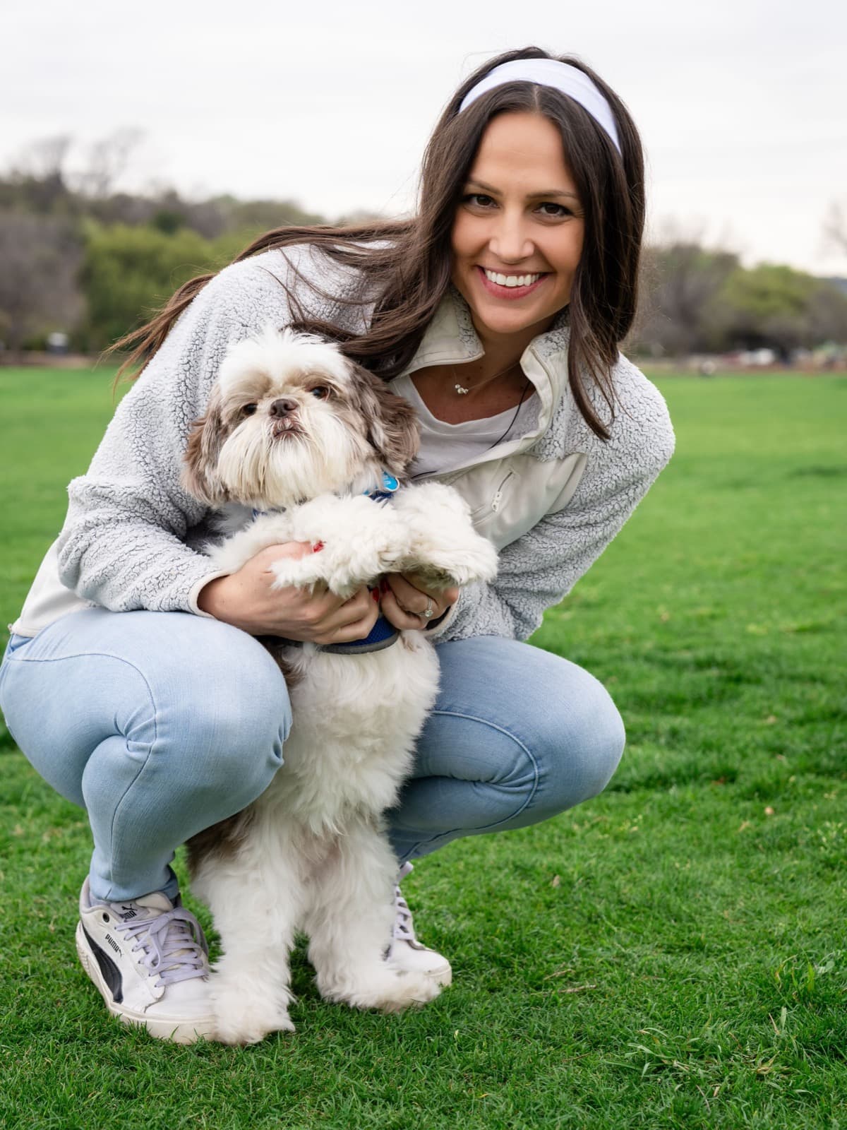 Tori Lee crouching on the grass in a park, smiling and holding a small white dog