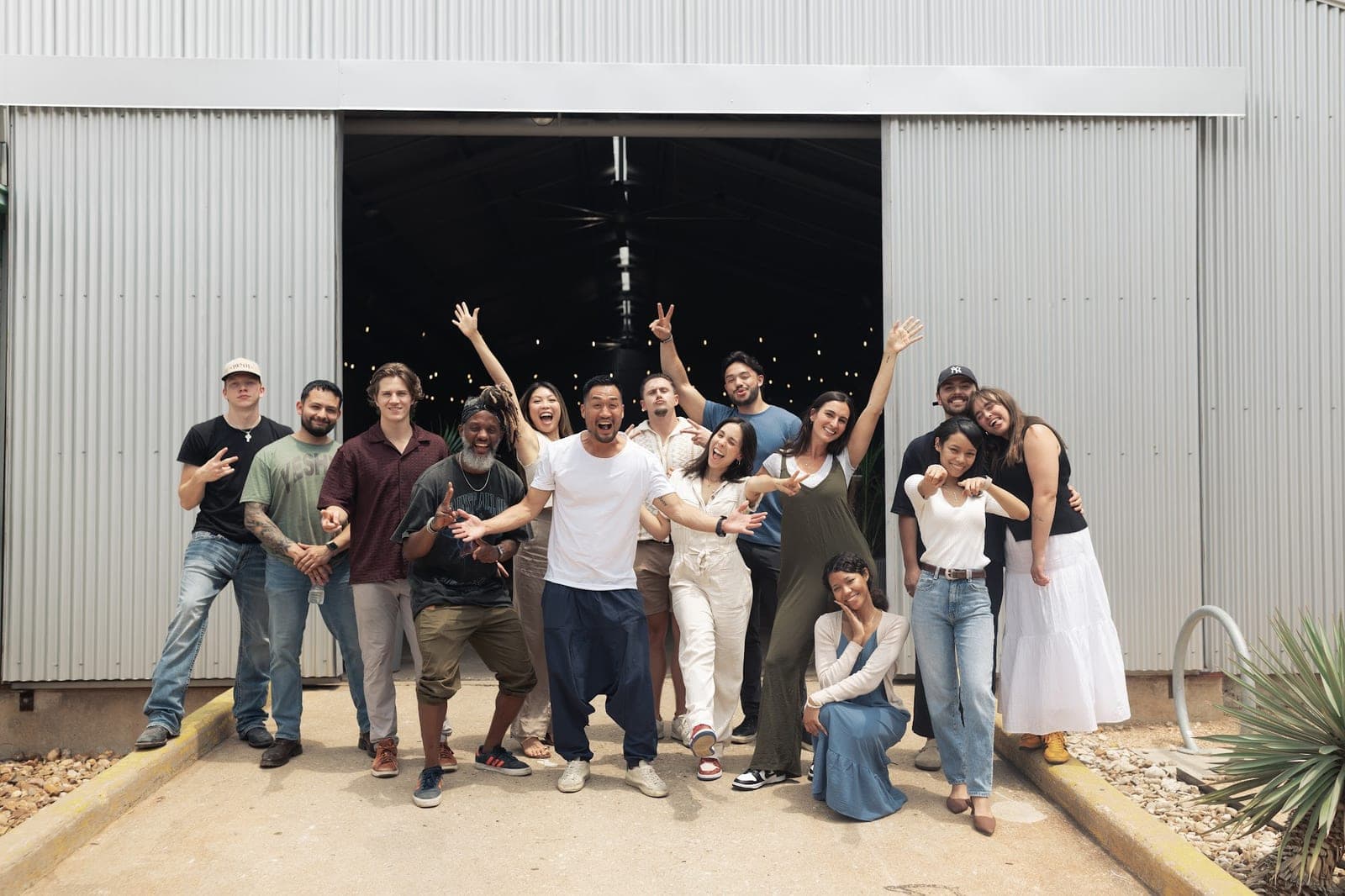 About fifteen people cheer and wave outside the open roll-up door of a corrugated metal building.