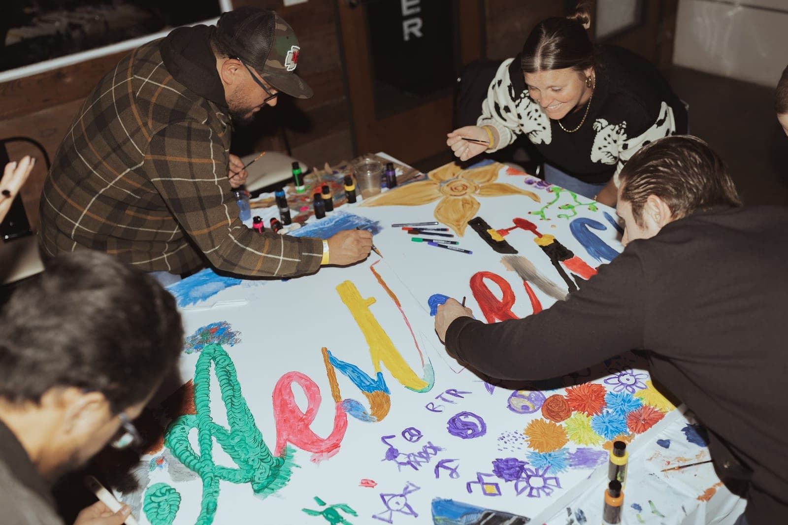 People lean over a long paper banner, painting the word Aesthetic in bright colors.