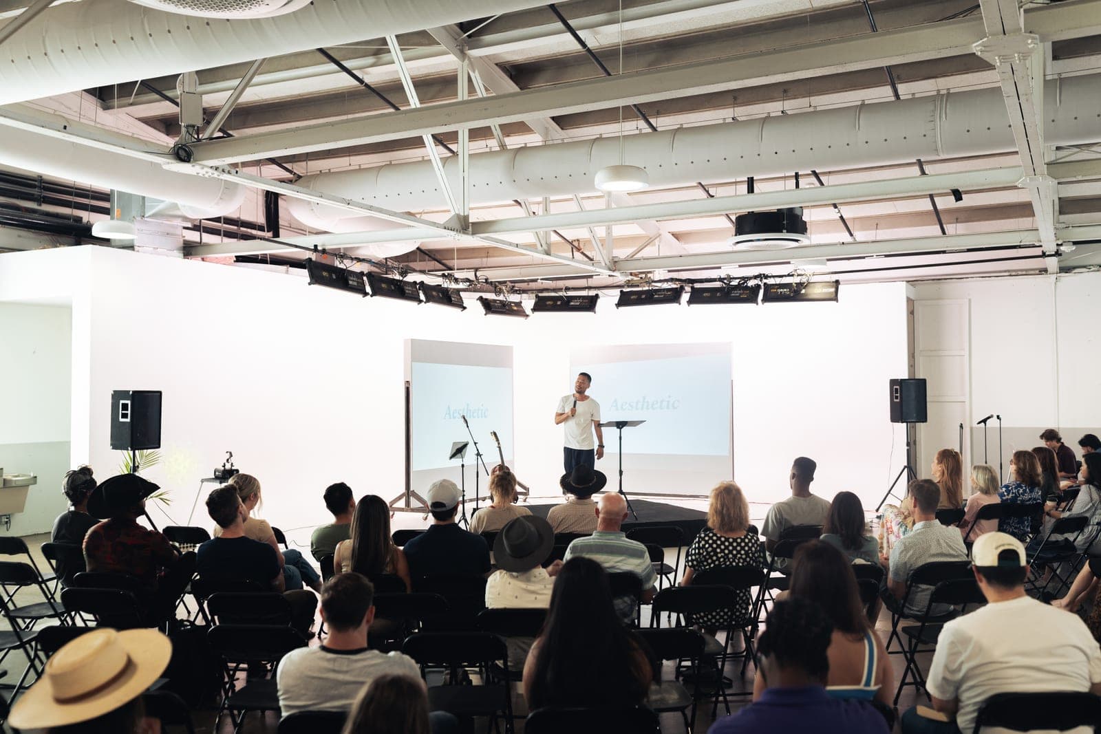 A speaker on a low stage between two screens reading Aesthetic, with the room seated in rows of folding chairs.